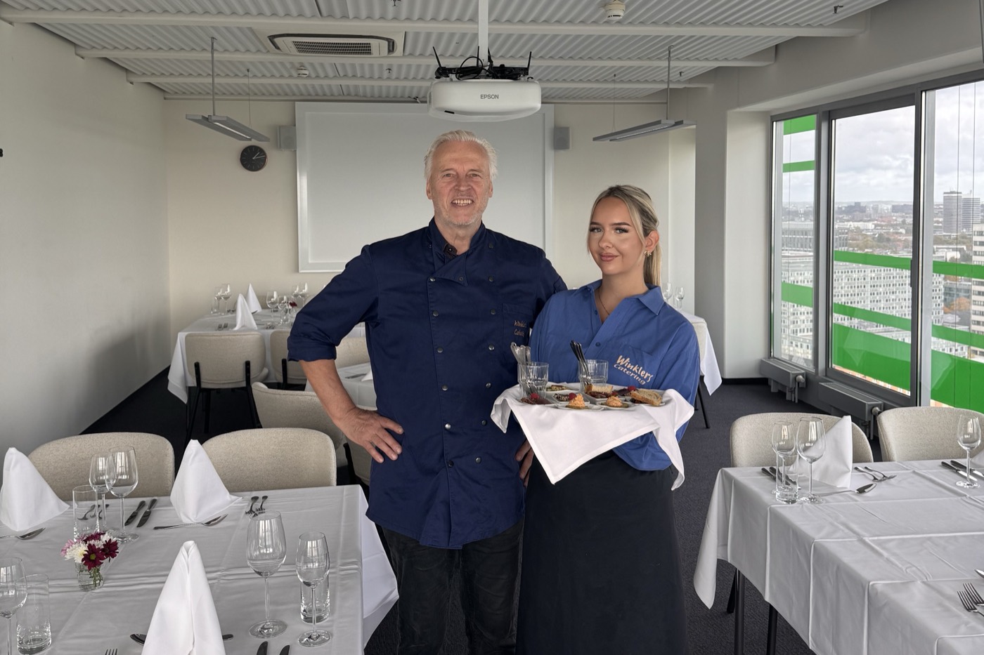 Servicekraft von TopS. mit Tablett neben dem Küchenchef in einem eingedeckten Veranstaltungsraum